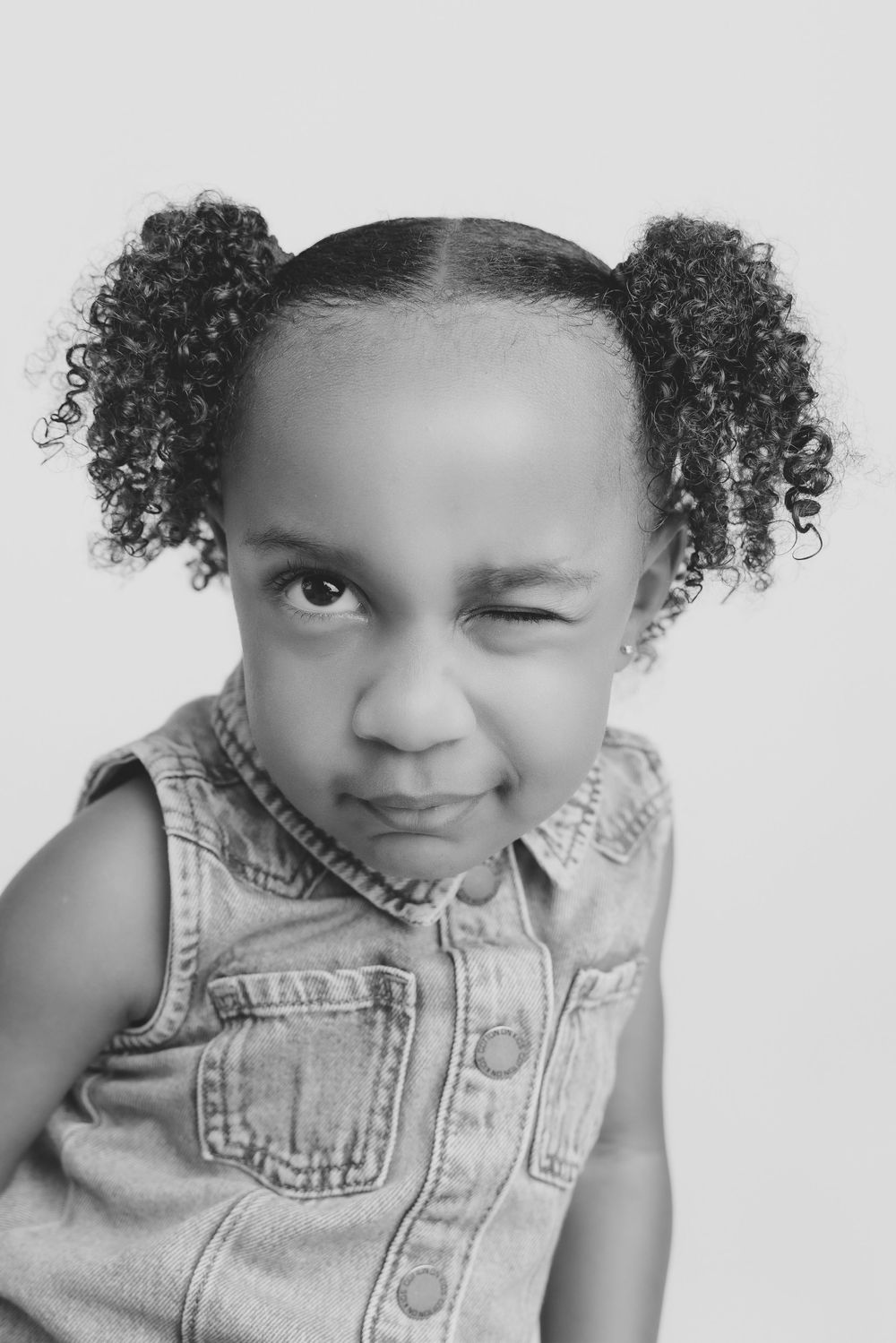 Black and white portrait of a young girl with curly pigtails wearing a denim vest, making a playful squinting expression.