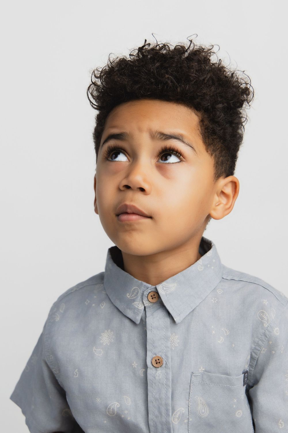 Young boy with curly hair wearing a gray button-up shirt, looking upward thoughtfully against a white background.