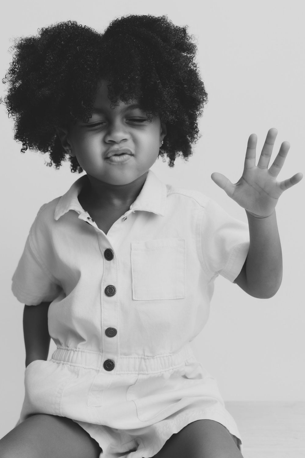 Black and white portrait of a young girl with curly afro hair, wearing a button-up romper, raising her hand.