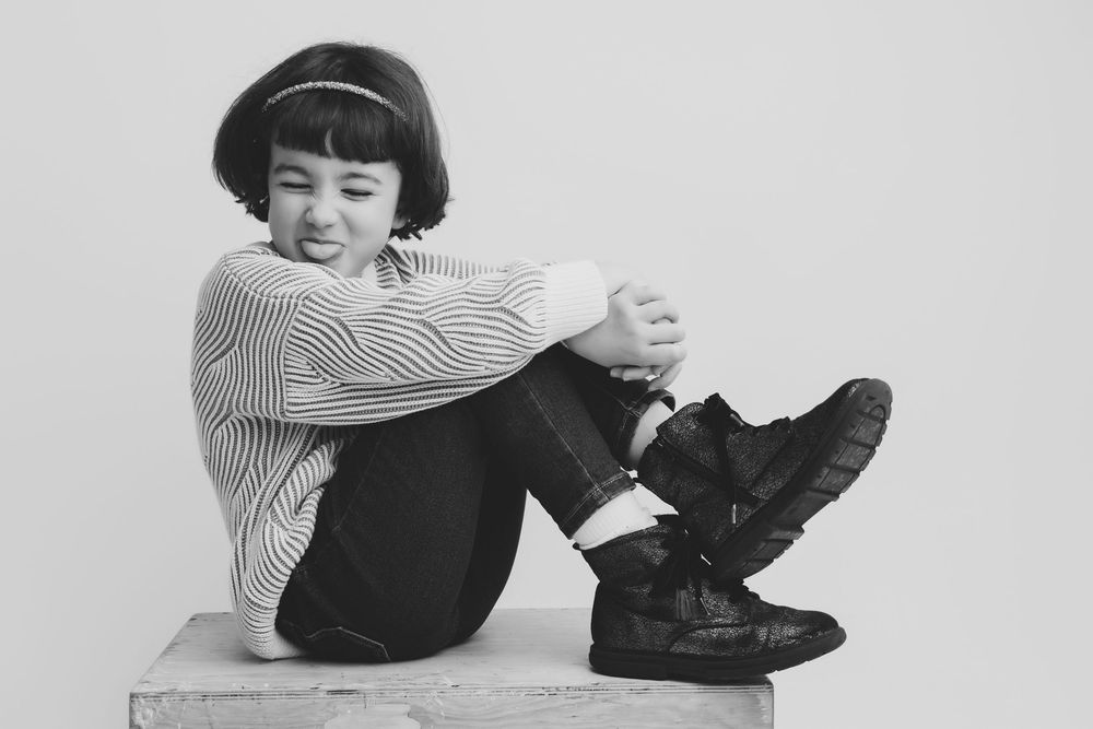Black and white photo of a young girl sitting on a block, wearing striped shirt and boots, hugging her knees.