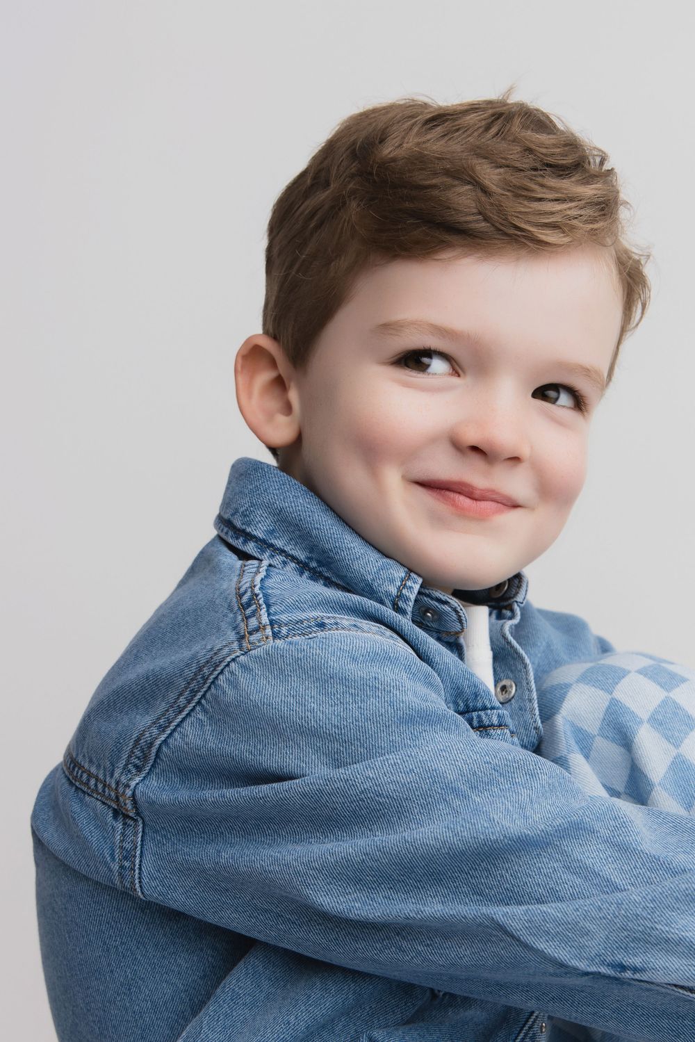 Smiling young boy in denim jacket and checkered shirt posing against light gray background.