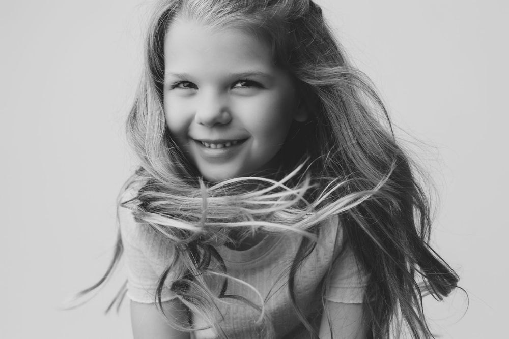 Black and white portrait of a smiling young girl with long flowing hair against a light background.