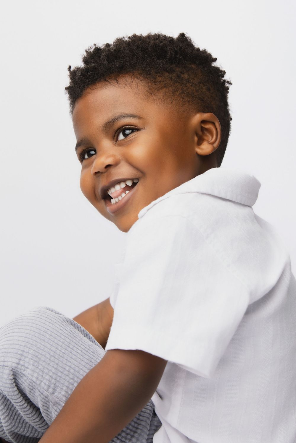 A cheerful young Black boy in a white polo shirt smiles over his shoulder against a light gray background.
