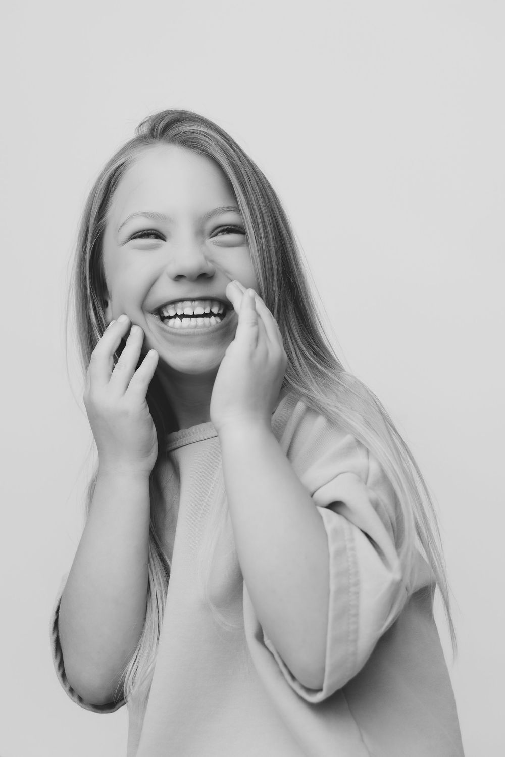 Black and white portrait of a laughing young girl with long hair, hands on cheeks, against light background.