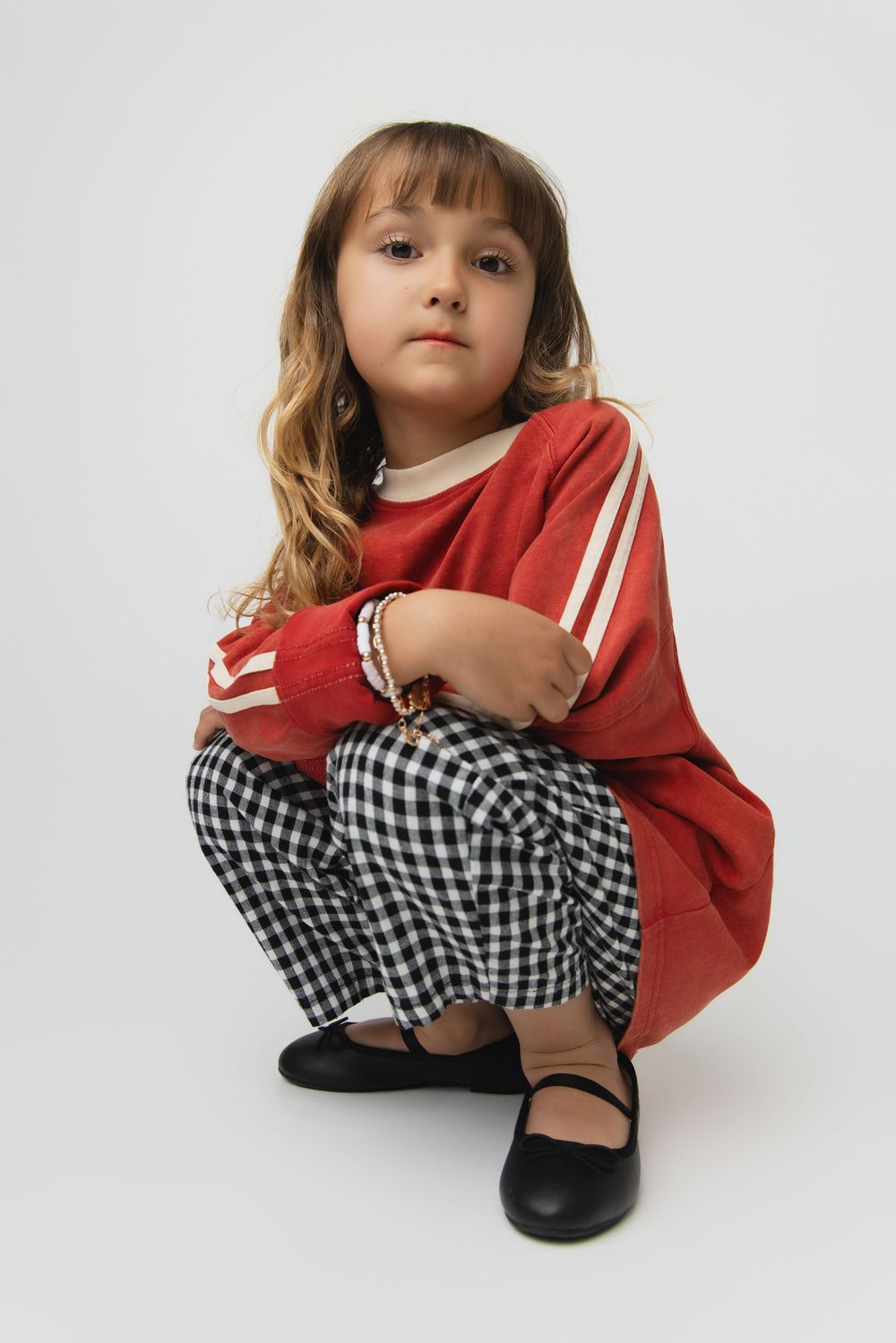 Young girl in red striped top, checkered pants, and black flats crouching on white background.
