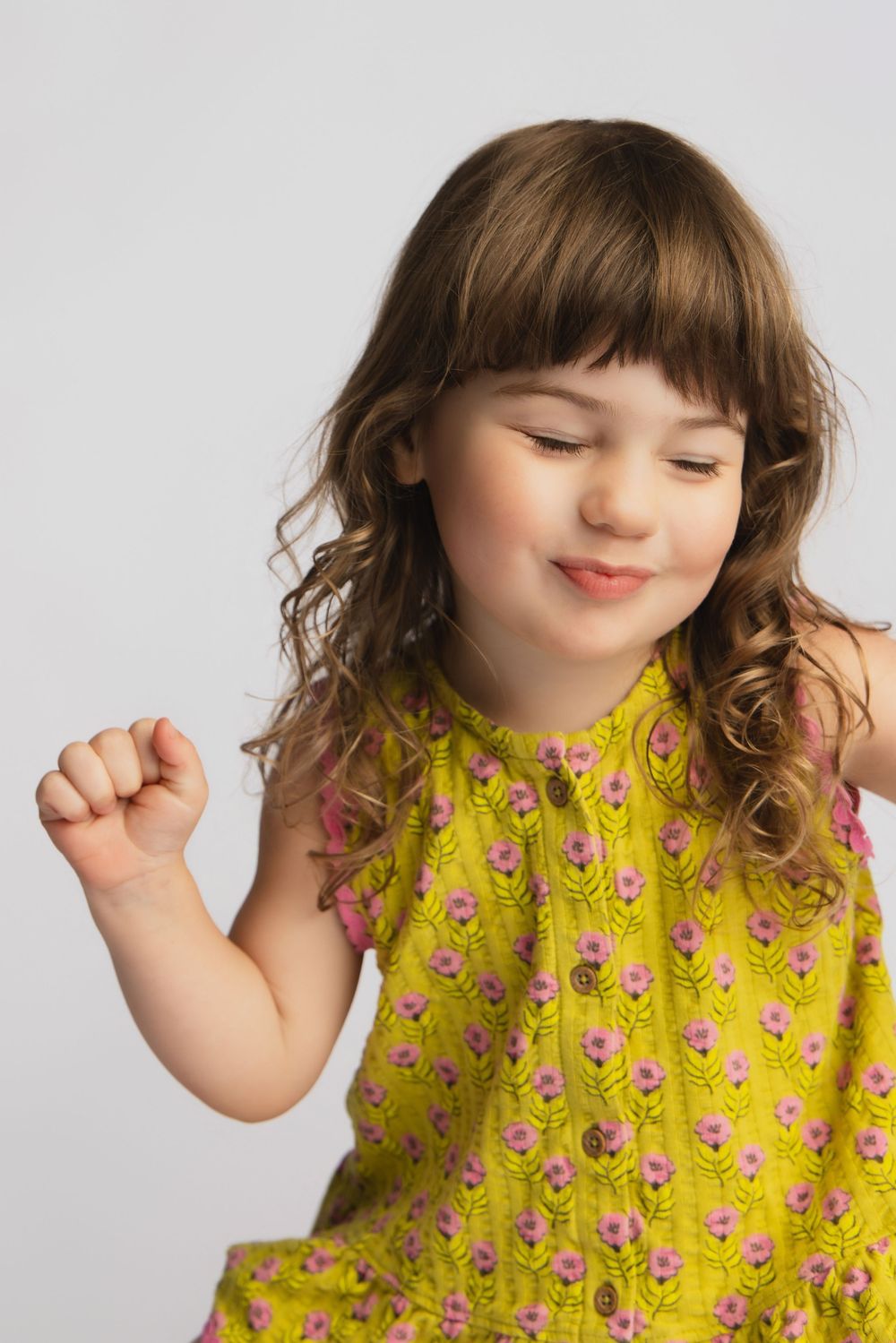 Young girl in yellow floral dress dancing with eyes closed, smiling joyfully against a white background.