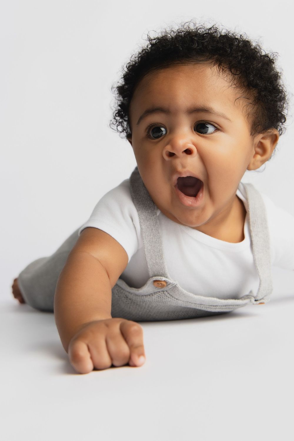 Adorable African American baby lying on stomach with mouth open in surprise, wearing white onesie and gray overalls.