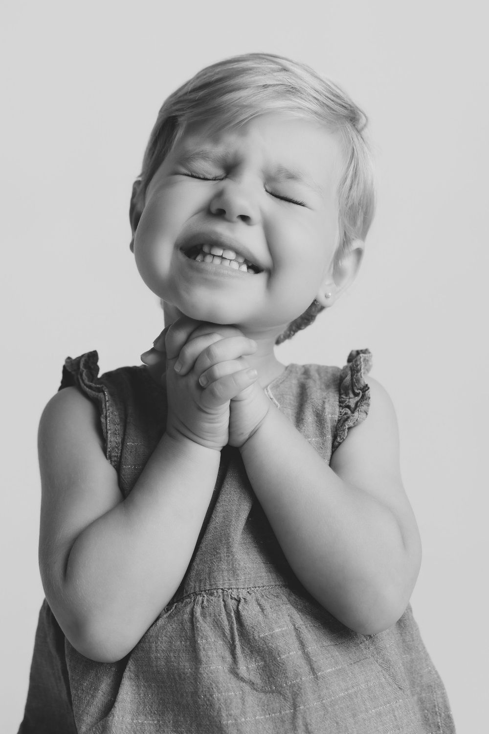 Black and white photo of a joyful toddler girl with clasped hands, eyes closed, smiling brightly in a sleeveless dress.