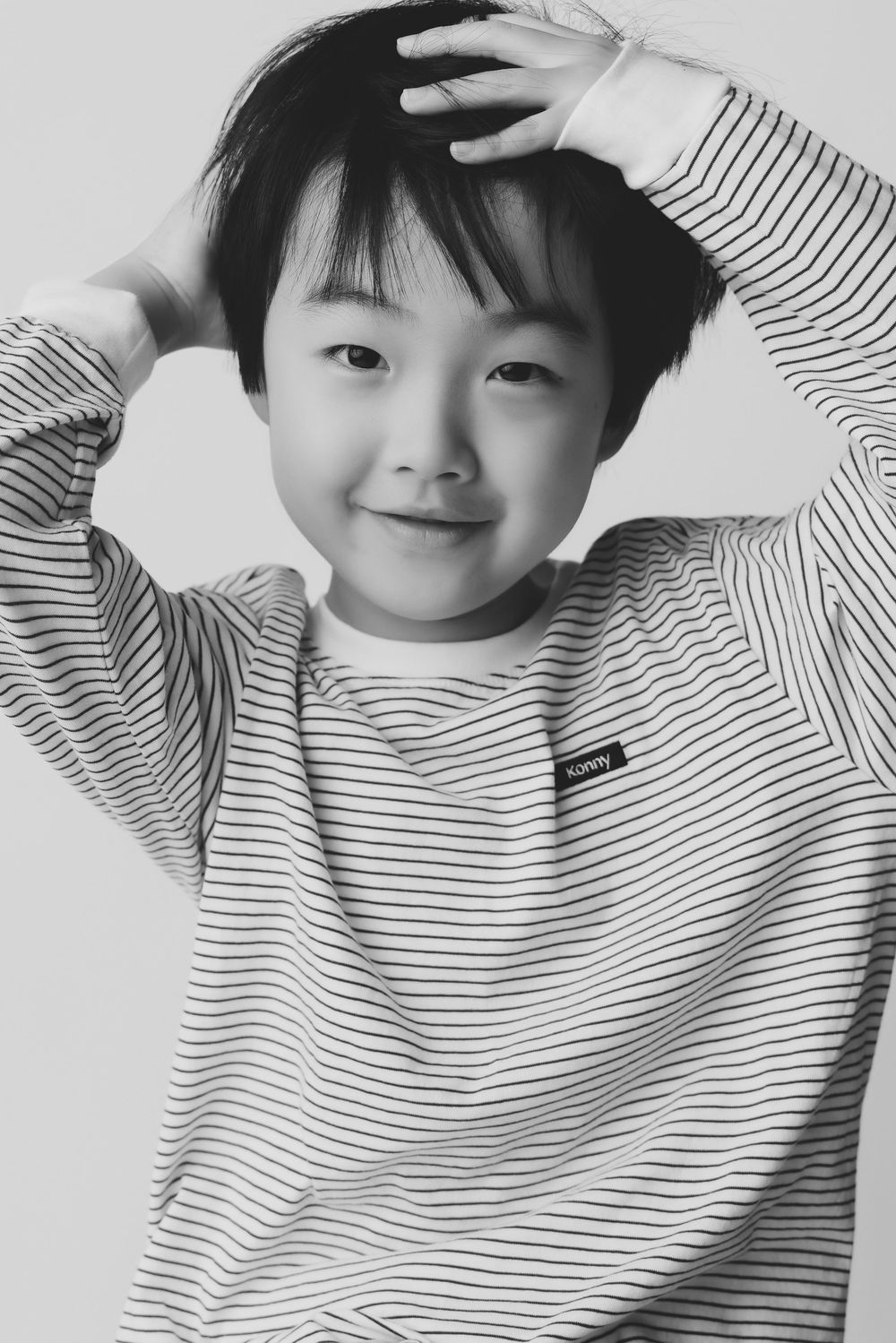 Black and white portrait of a smiling Asian boy in a striped sweatshirt with hands raised behind his head.