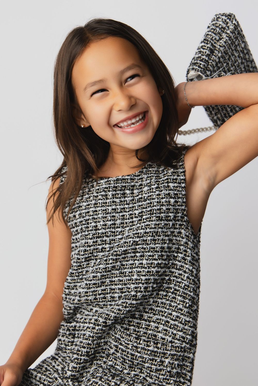 Smiling young girl in a black and white tweed sleeveless dress, holding a small purse behind her head.