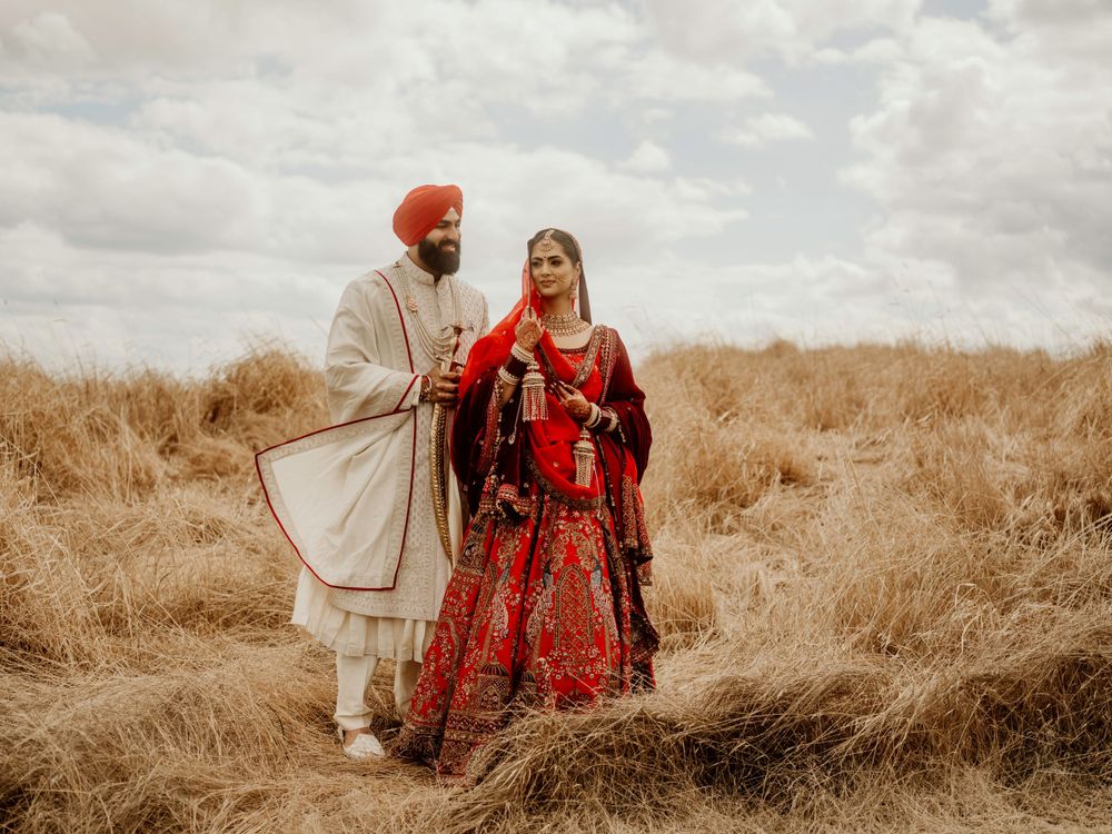 A couple in traditional Indian wedding attire stand together in a golden grass field under cloudy skies.