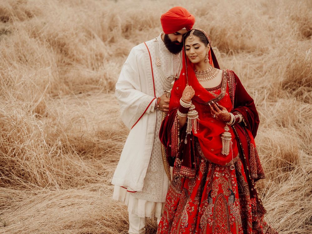 A couple in traditional Indian wedding attire poses in a field of tall dried grass with warm sepia tones.
