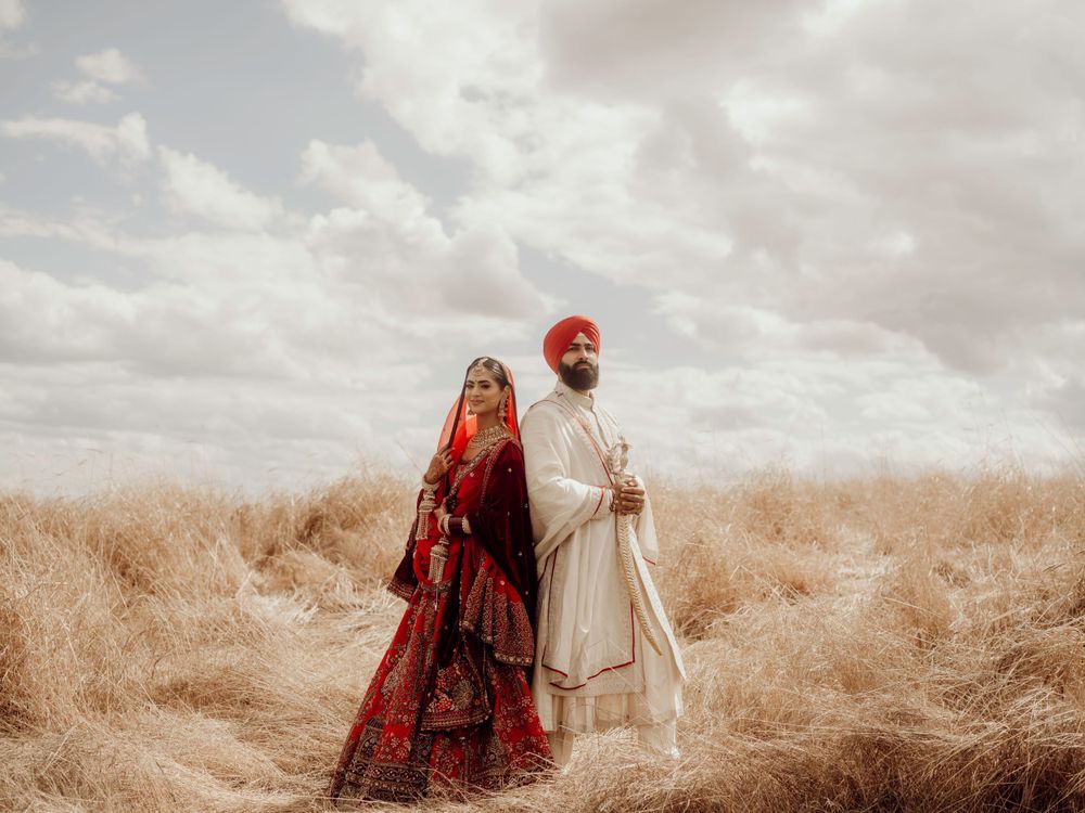 Couple in traditional Indian wedding attire stands together in a golden wheat field under cloudy sky.