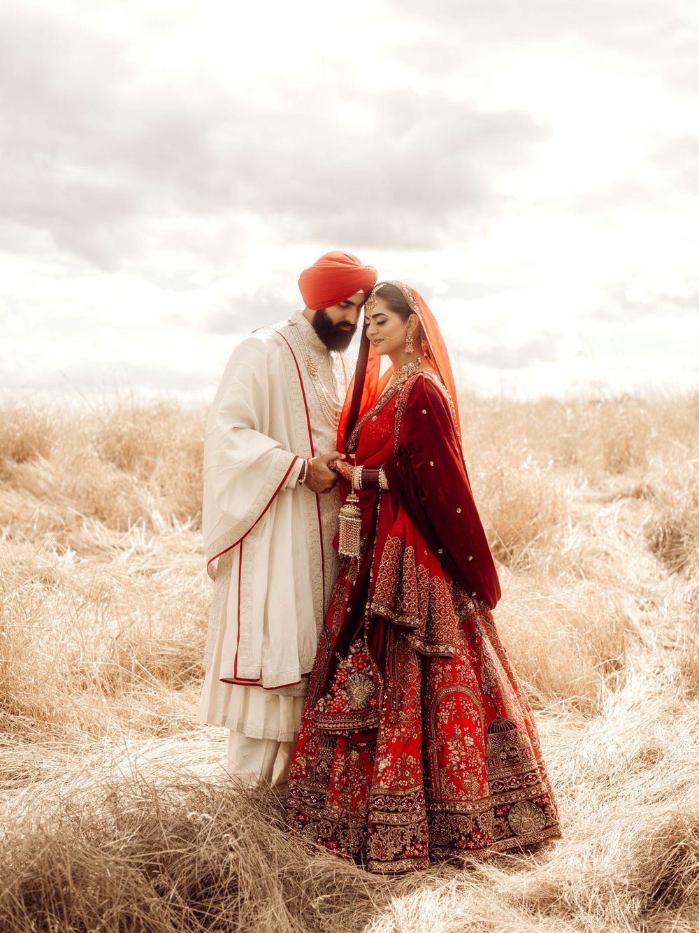 A couple in traditional Indian wedding attire embrace in a dreamy sepia-toned field wearing red and cream ceremonial clothing.