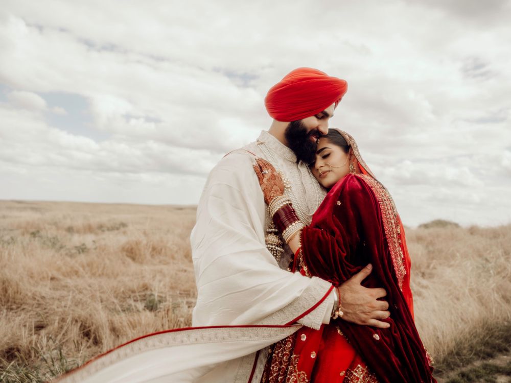 A couple in traditional Indian wedding attire embrace in a grassy field under cloudy skies.