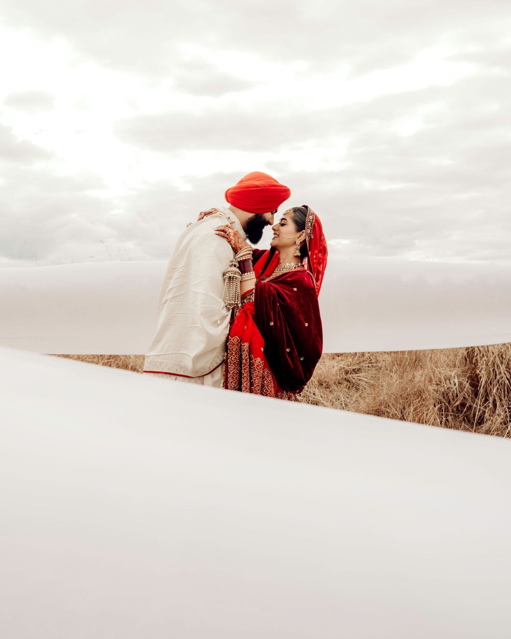 A couple in traditional Indian attire embraces on a snowy hillside against an overcast sky.