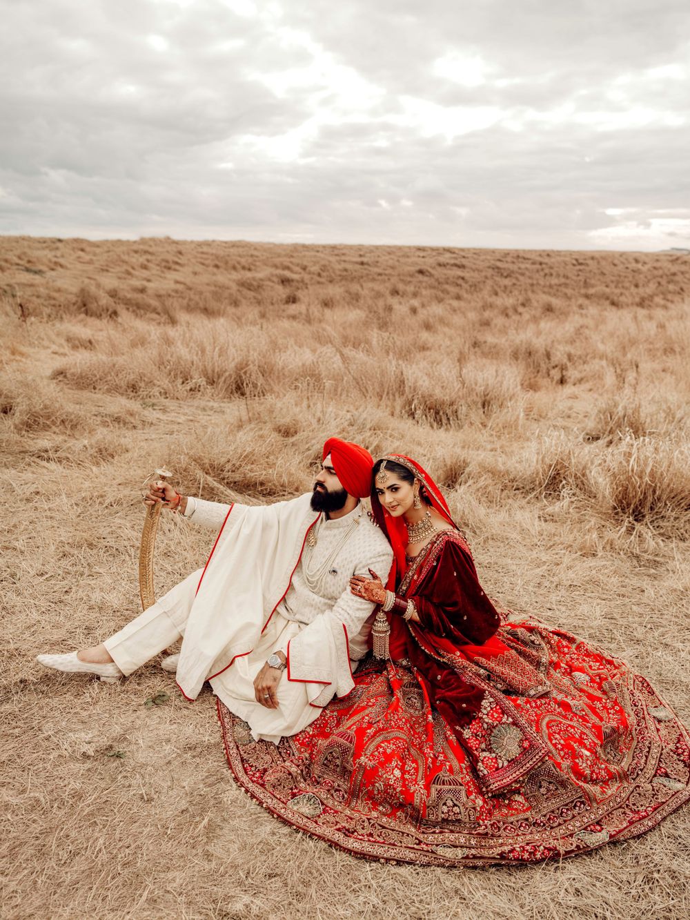 A couple in traditional Indian wedding attire posing in a desert landscape with red lehenga and cream sherwani outfits.