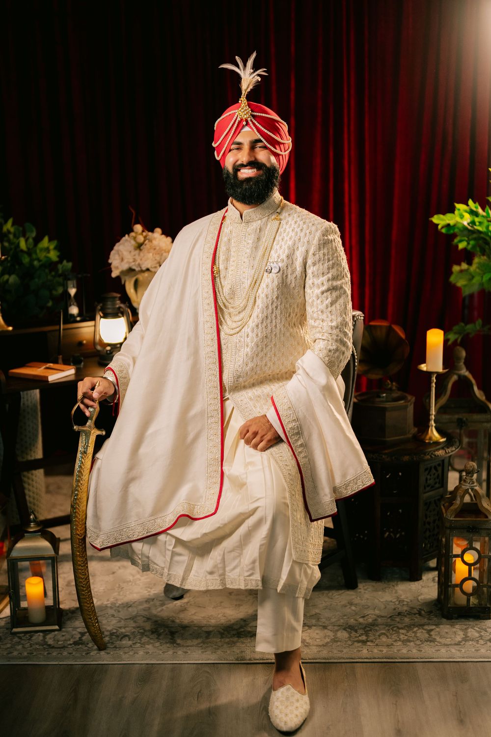 Traditional Indian groom wearing elegant white sherwani, red turban, and beaded necklace poses with a cane near lanterns.