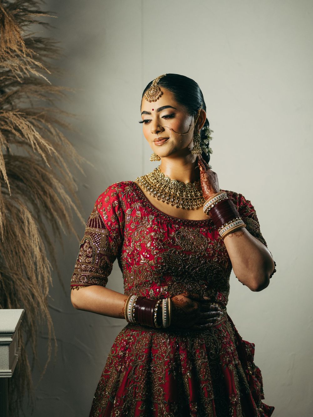 Traditional bride in ornate red lehenga with gold jewelry and embellishments poses elegantly against pampas grass decor.