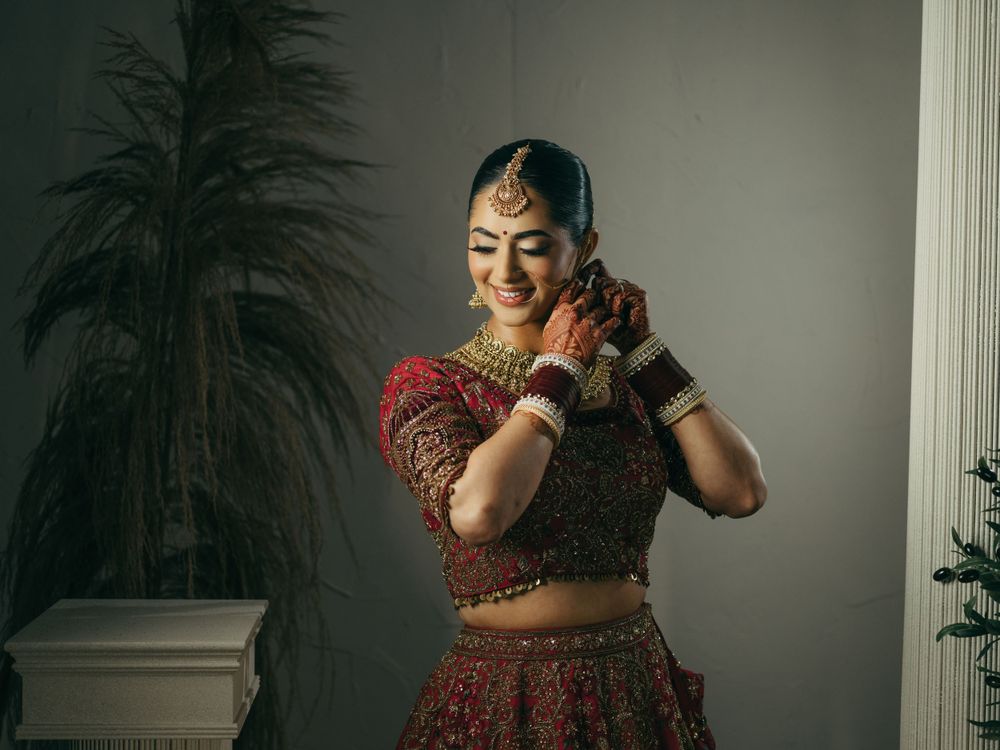 A graceful woman in traditional Indian bridal attire with a red lehenga adjusts her earrings while smiling.