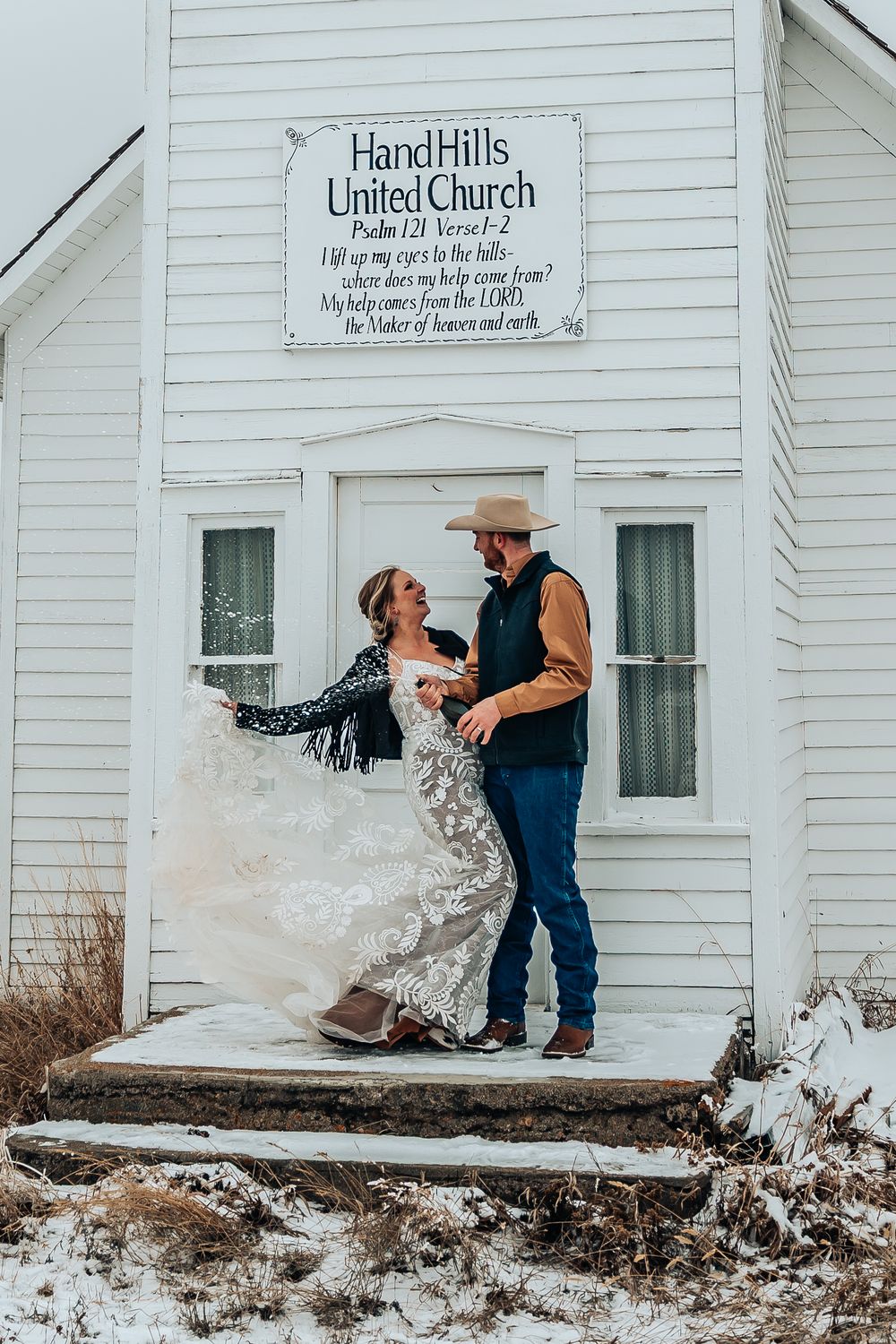 Romantic scene outside HandHills United Church with couple in western and bridal attire.