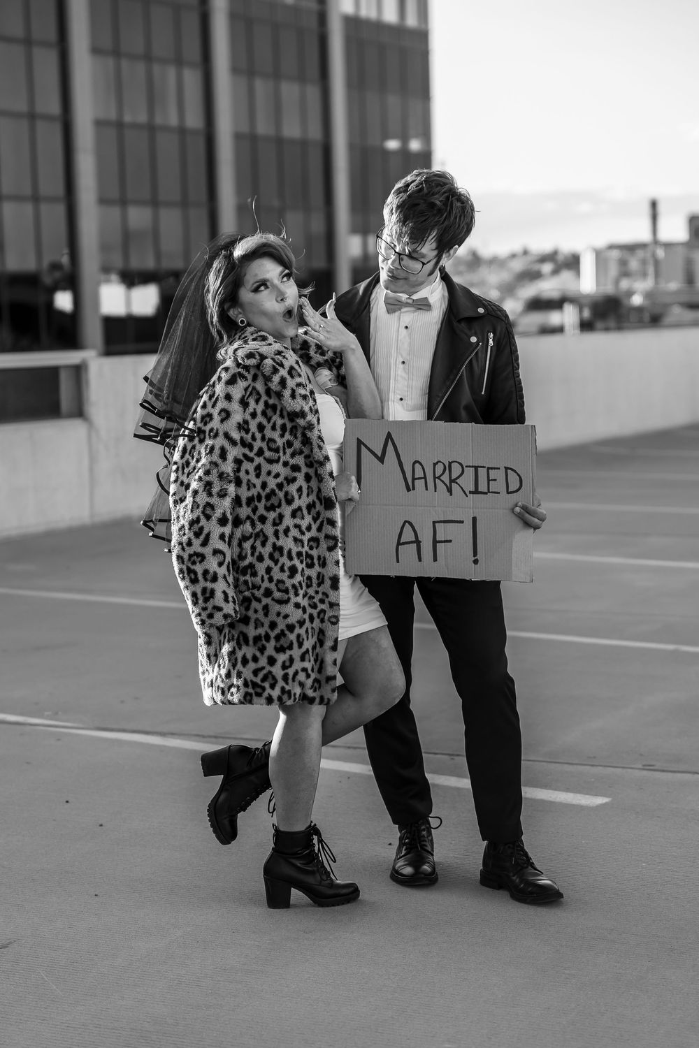 Couple poses with 'Married AF' sign, bride wearing leopard print coat in parking structure.