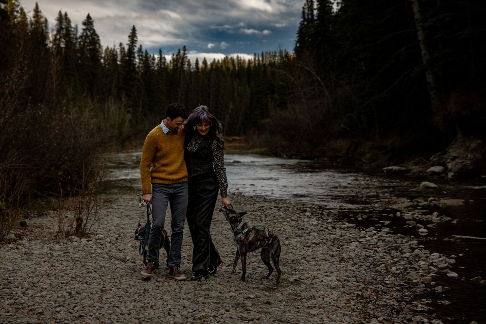 Two people and their dog stand together on a rocky riverbank surrounded by evergreen forest on a cloudy evening.