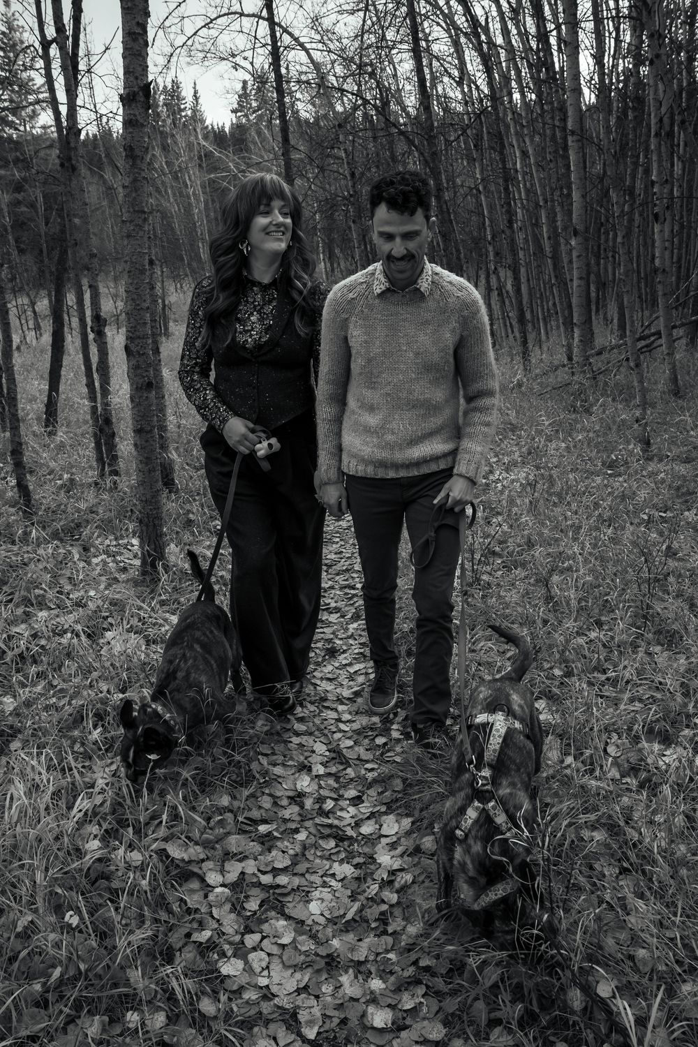 A couple walks together through a wooded forest trail covered in fallen autumn leaves.