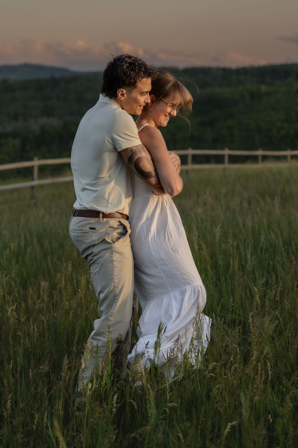 A romantic couple embraces in a field with rolling hills and golden light in the background.