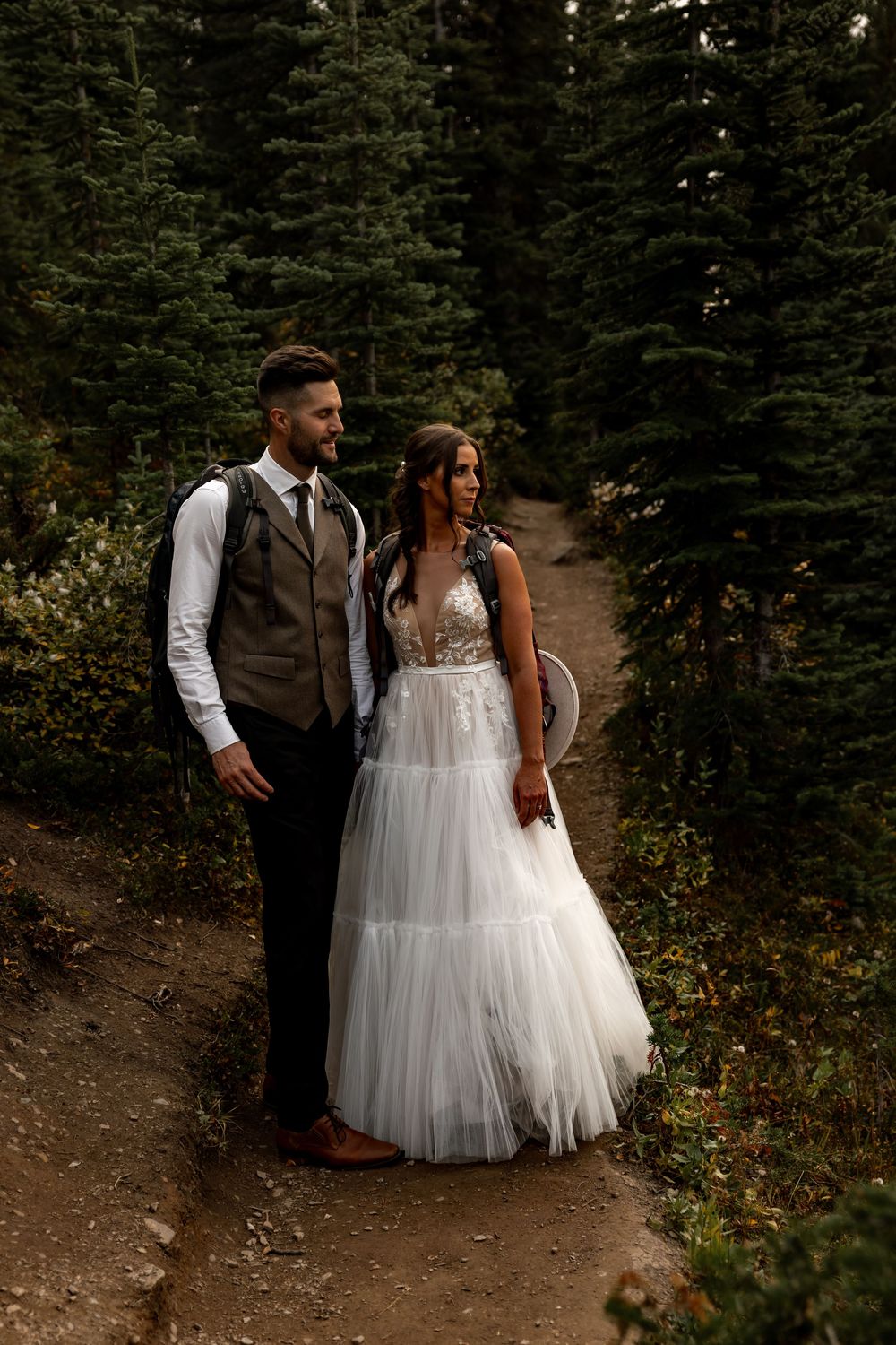 A couple in wedding attire walks along a forest trail surrounded by evergreen trees.