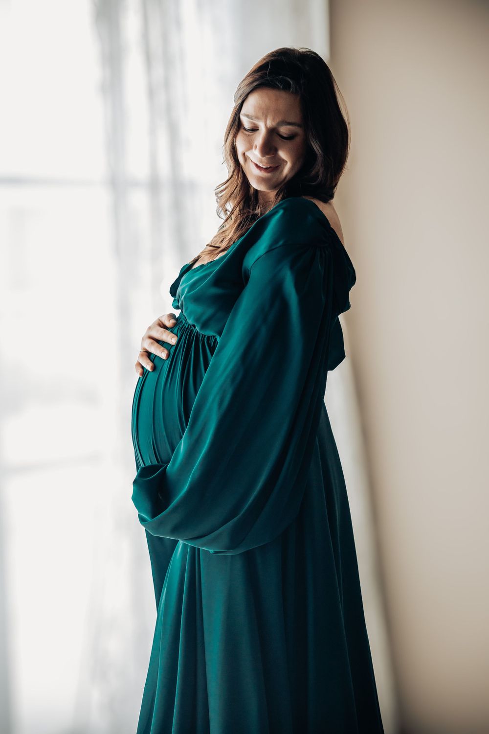 Elegant poses in a flowing deep green dress captured near bright windows.