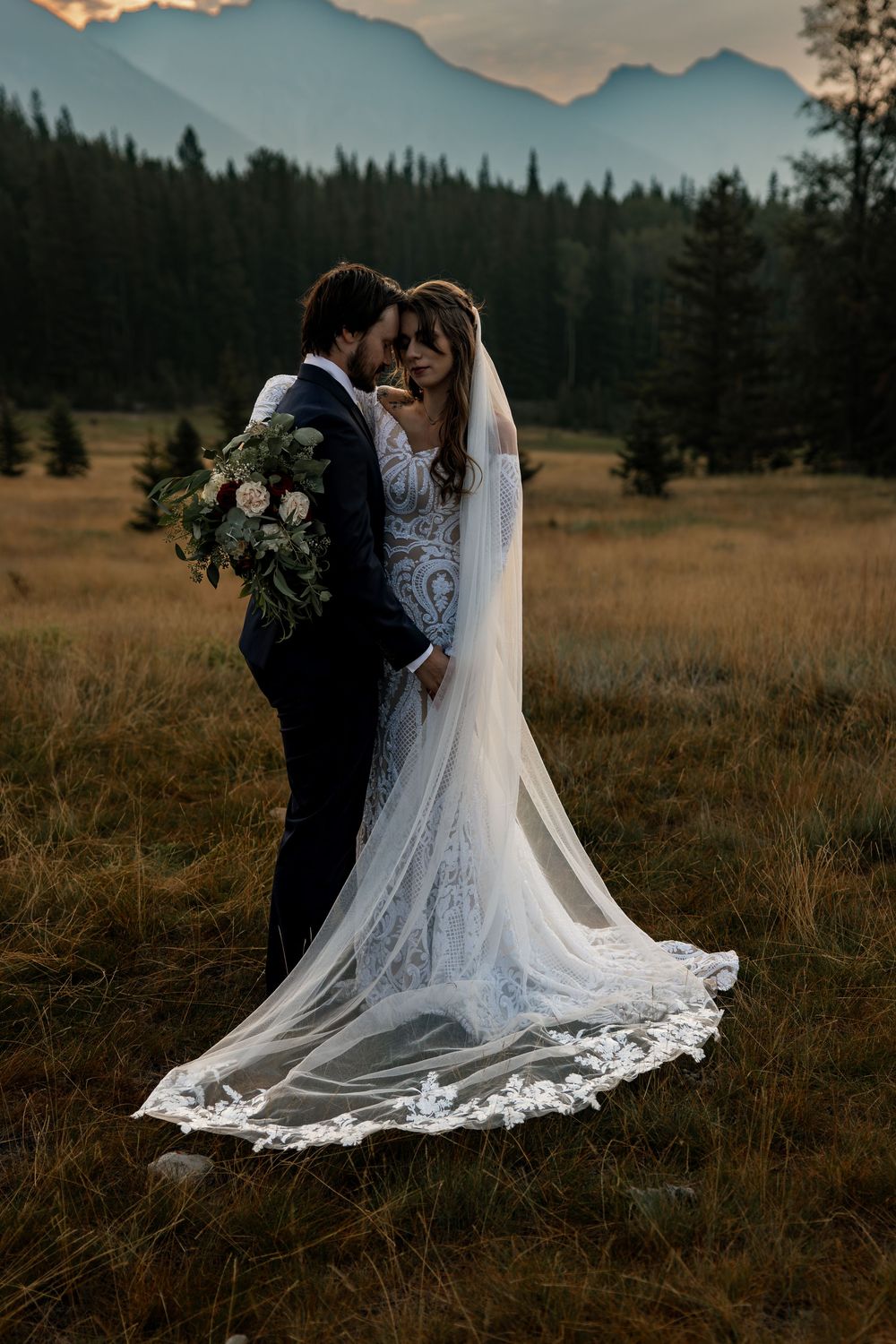 A romantic wedding couple stands in a golden meadow with mountains and evergreen forest in the background at sunset.