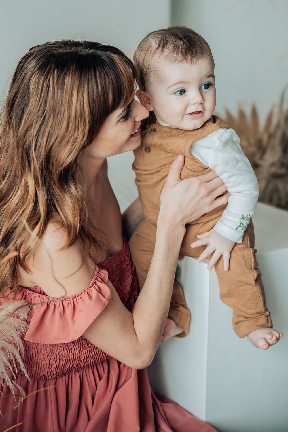 Parent in coral dress shares intimate moments with baby against light wall and dried flowers.