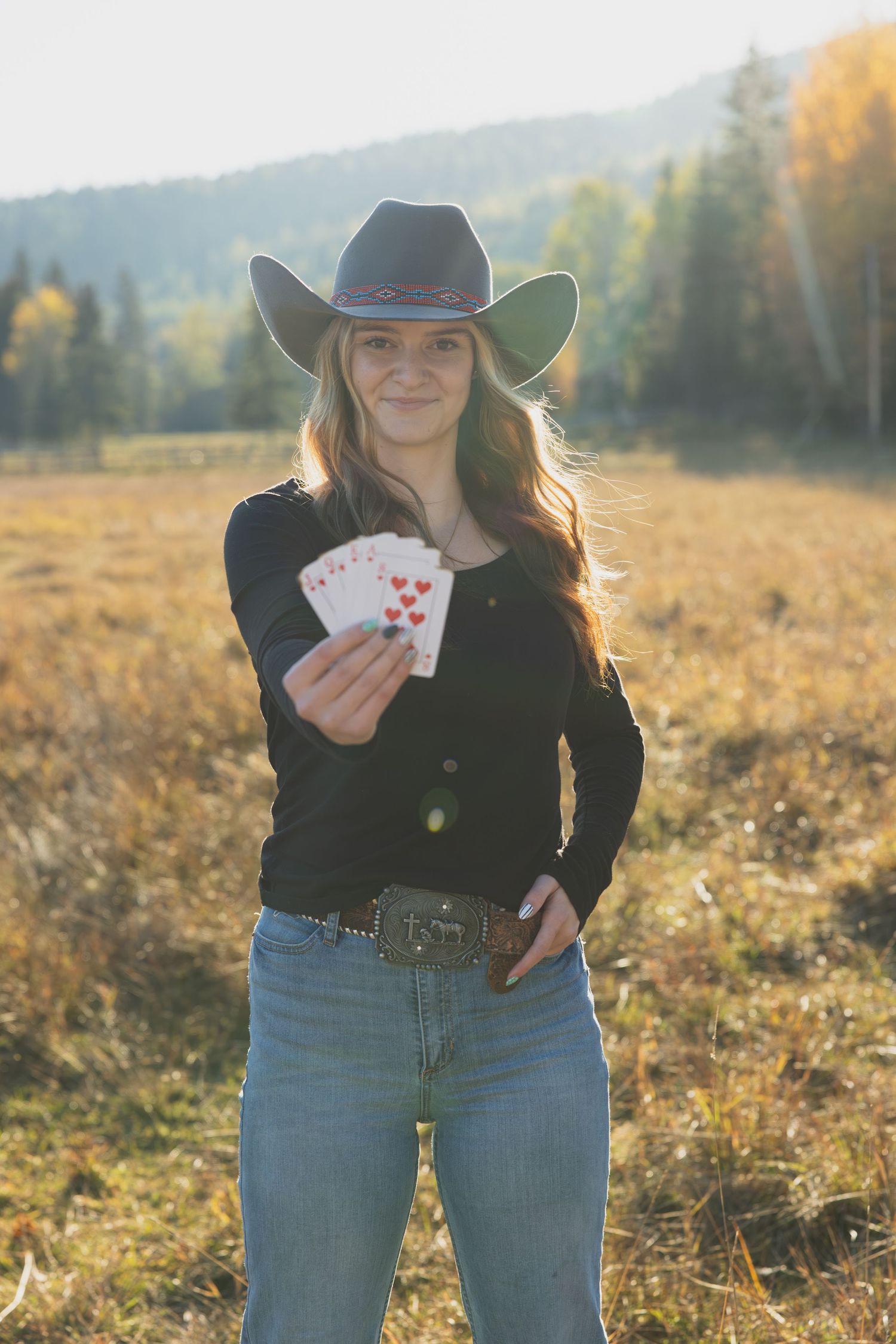 Smiling young woman in cowboy hat and jeans holds playing cards in a sunny autumn field.