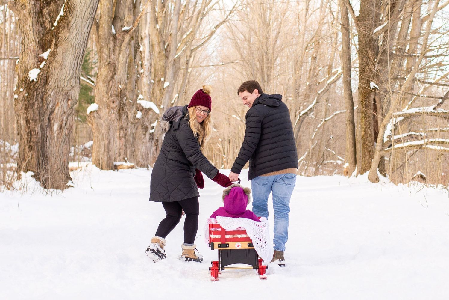 Family pulling a bundled toddler on a red sled through a snowy winter forest path.