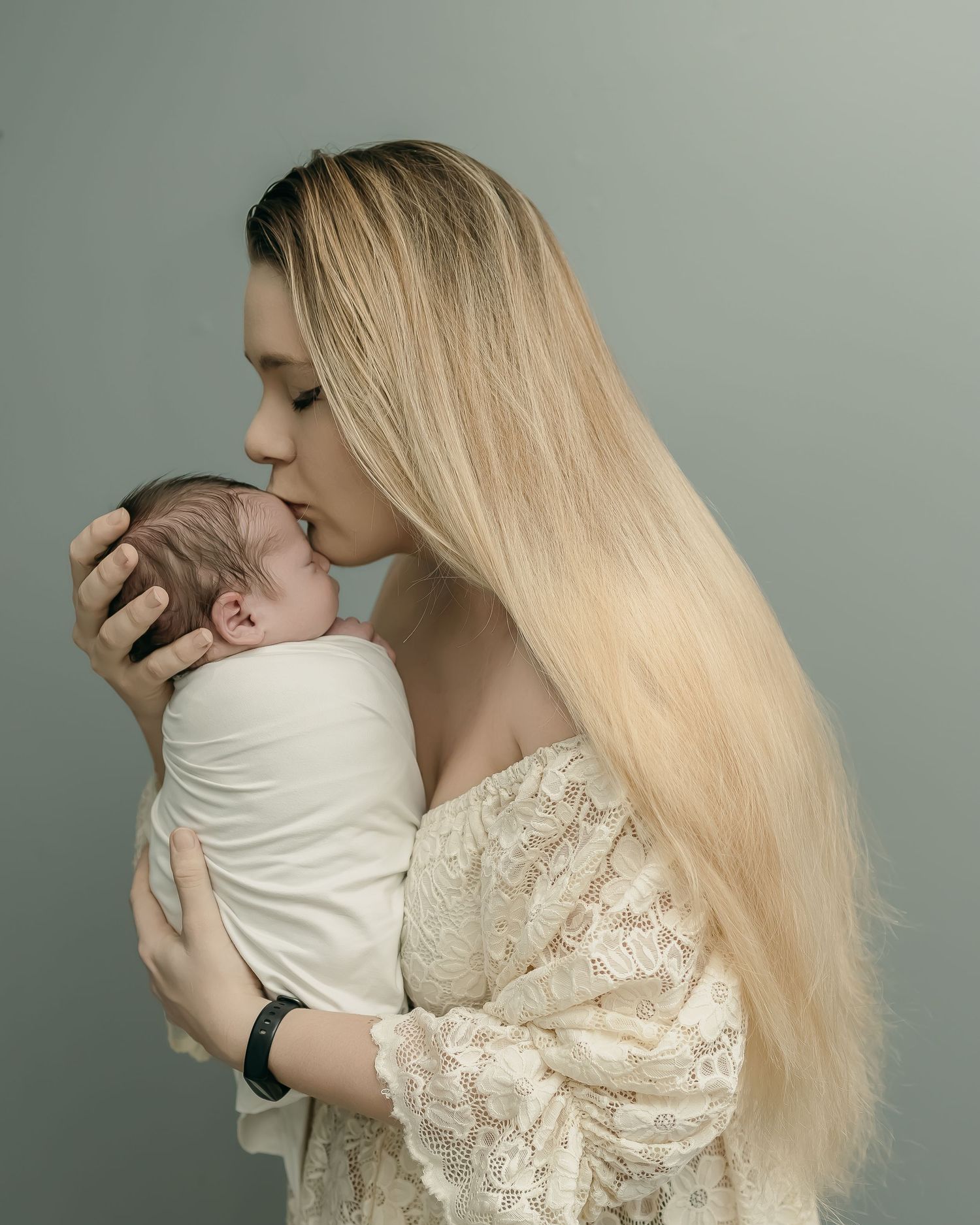 A tender moment between a woman with long blonde hair in a lace dress holding and kissing a swaddled newborn baby.