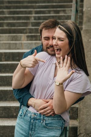 A joyful couple embracing on concrete stairs shows excitement while displaying an engagement ring during a proposal at Piedmont Park Atlanta