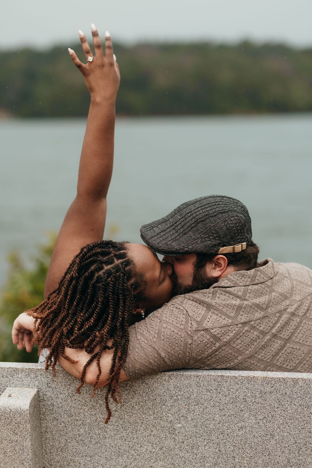 A person with dreadlocks wearing a newsboy cap lies on a concrete surface near a lake.