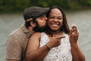A joyful couple shares a candid moment by the lake, with one kissing the other's cheek as they laugh together.