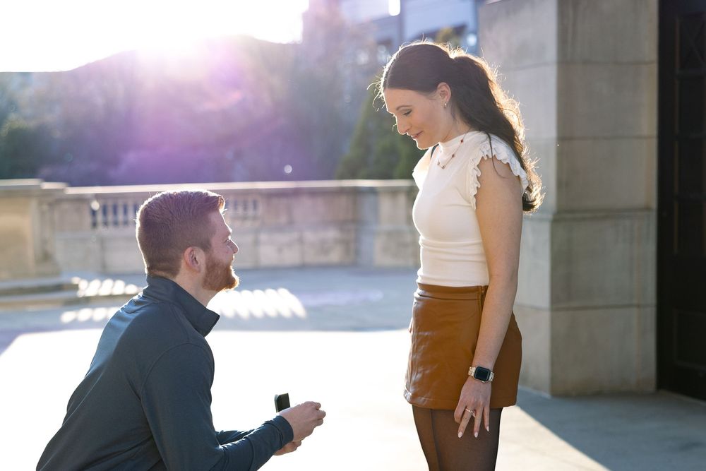 Man proposing on one knee to woman outdoors, holding ring box, with sunlight glowing behind them.