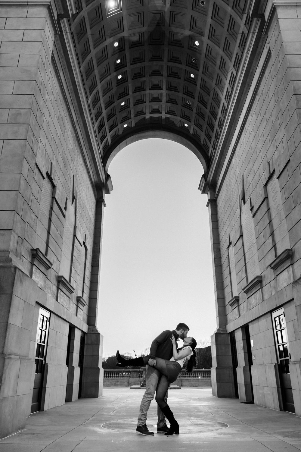 Black and white photo of a couple sharing a romantic dip kiss under a grand stone arch at dusk.