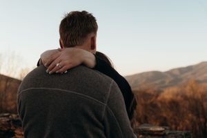 A couple embraces on a mountain overlook during sunset during a proposal showing her ring.