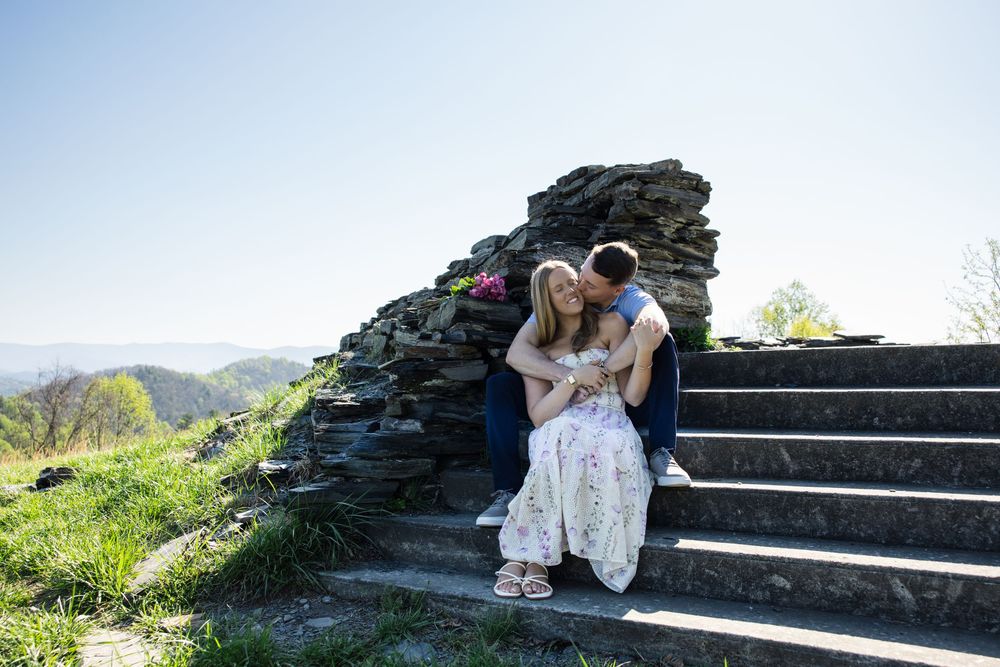 Couple sitting on stone steps outdoors, embracing romantically with scenic green hills and clear blue sky behind them.