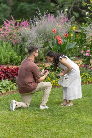 Man proposes on one knee to woman in white dress in a colorful garden, surrounded by blooming flowers.