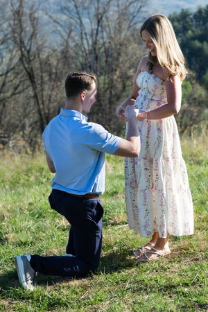 Man kneeling before pregnant woman in floral dress outdoors, holding tiny baby shoes in a sunny green field.