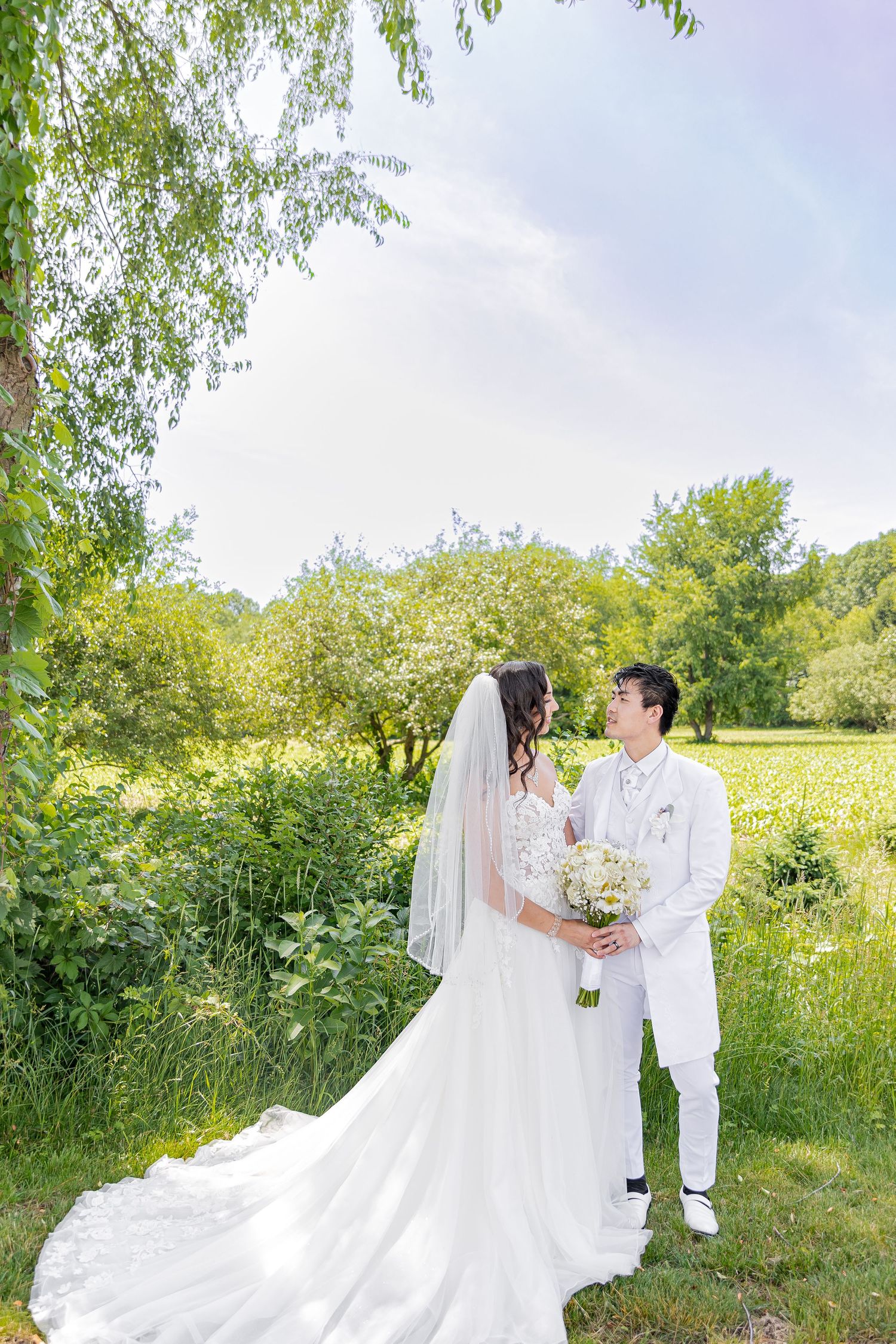 A wedding couple poses in a sunny meadow with flowing white dress and spring greenery.