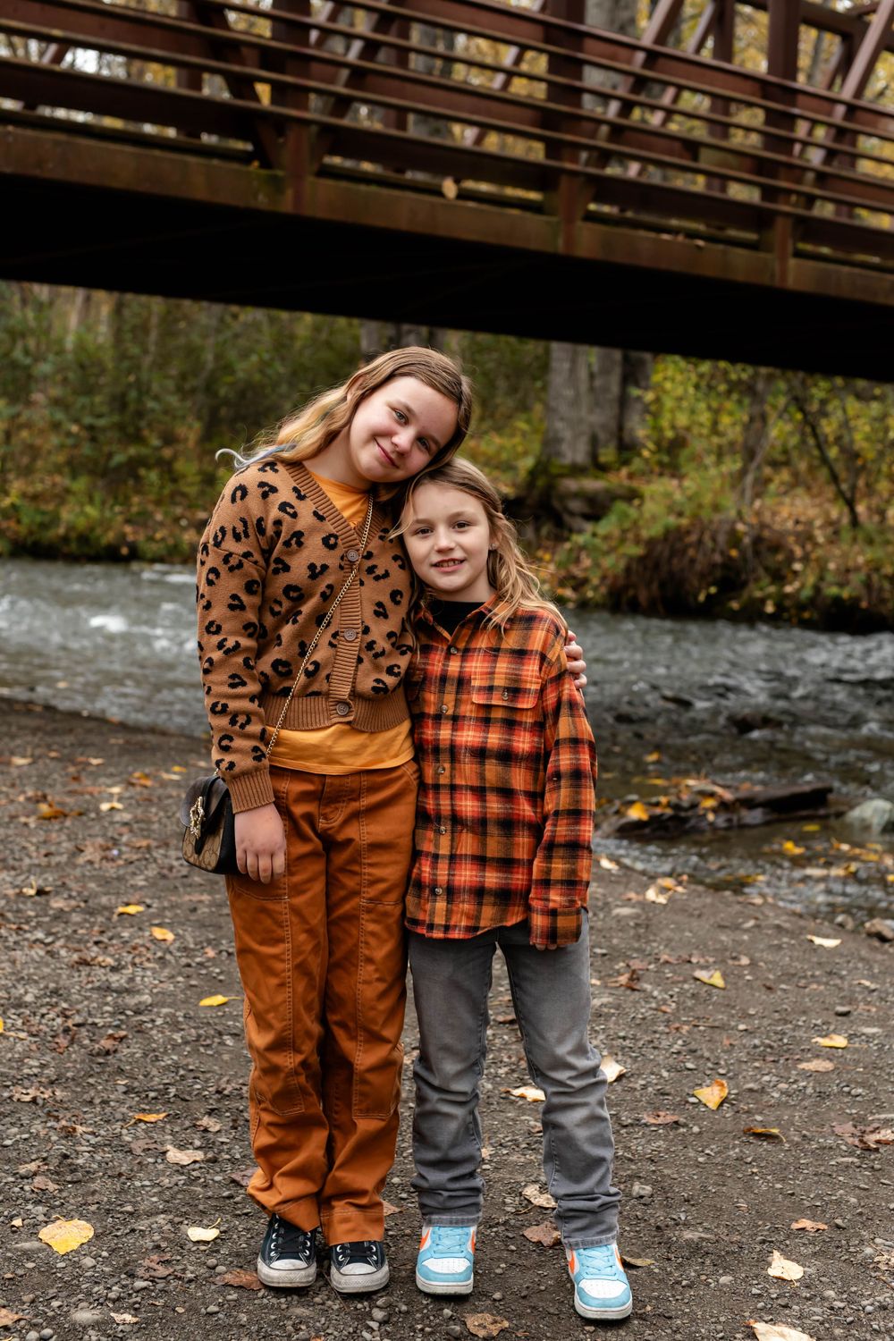 Two young people in autumn outfits pose in front of a wooden bridge and stream wearing orange and plaid patterns.