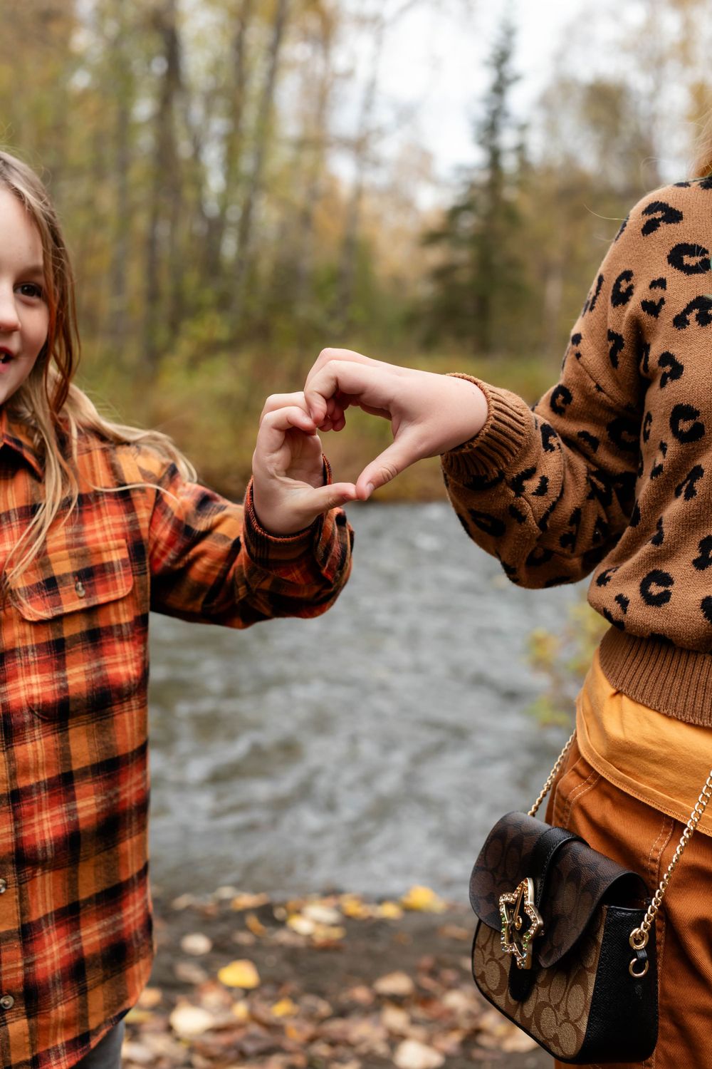Two people making heart shapes with their hands near a stream while wearing fall-themed clothing.