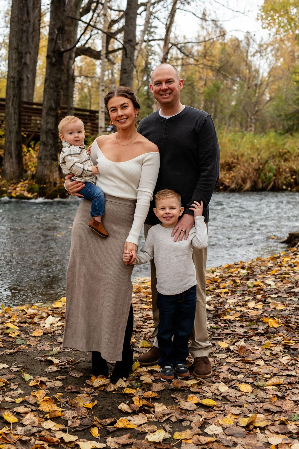 Family portrait by river with autumn leaves scattered on ground.