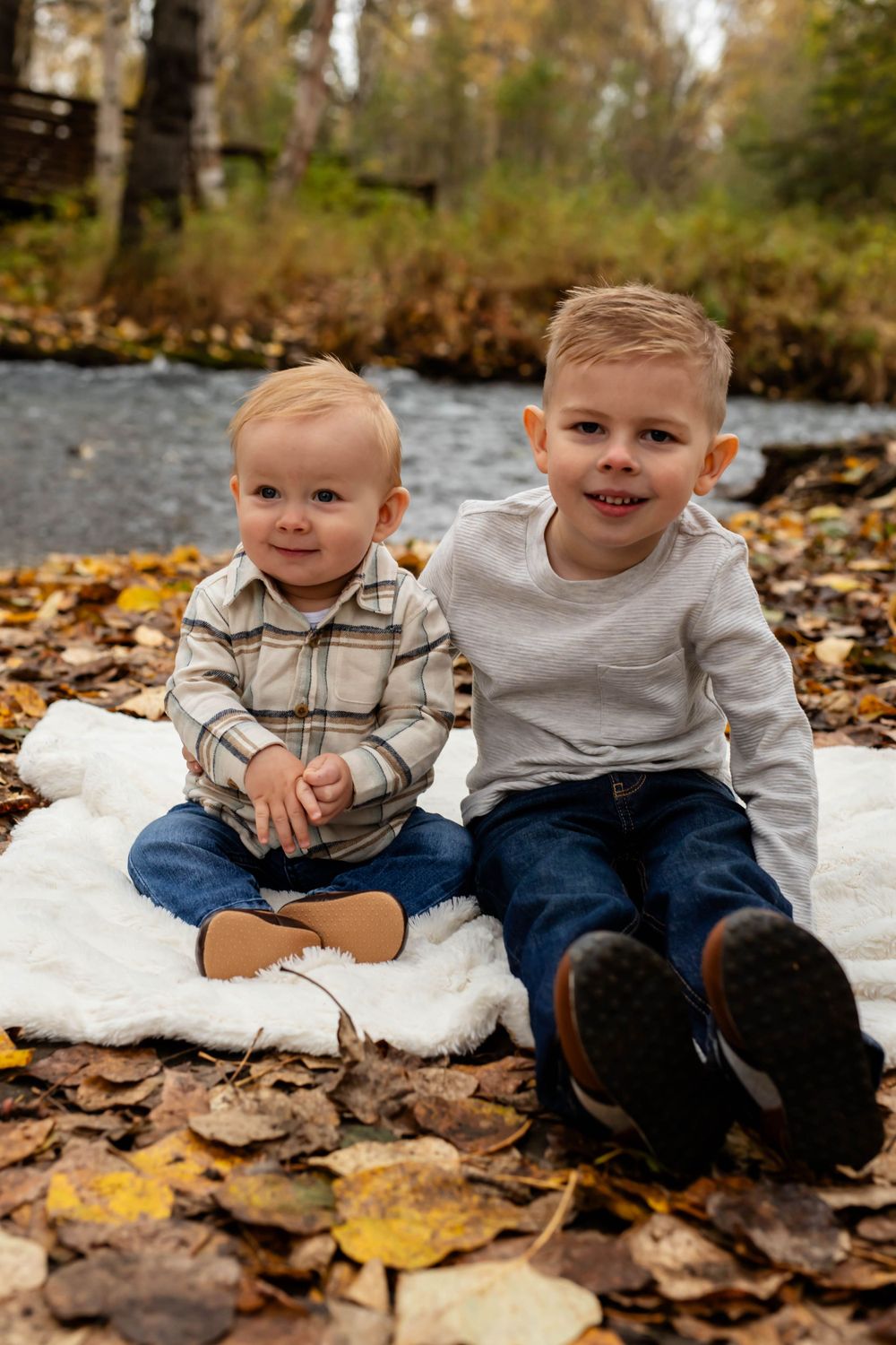 Two children sitting on white blanket among fallen autumn leaves.